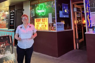 A night photograph of storytelling contest winner Molly McCloy holding her prize envelope outside The Brotherhood Lounge. The tavern is lit with vibrant red and green neon signs, and a window marquee reads "Story Oly! Heartbreak Tues June 16."