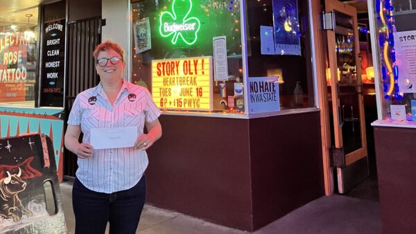 A night photograph of storytelling contest winner Molly McCloy holding her prize envelope outside The Brotherhood Lounge. The tavern is lit with vibrant red and green neon signs, and a window marquee reads "Story Oly! Heartbreak Tues June 16."